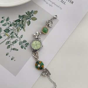 May include: A silver-toned wristwatch with a light green face and a decorative band. The band features floral and leaf accents, including a green and gold flower with a pearl center. The watch is on a white surface with text in the background.