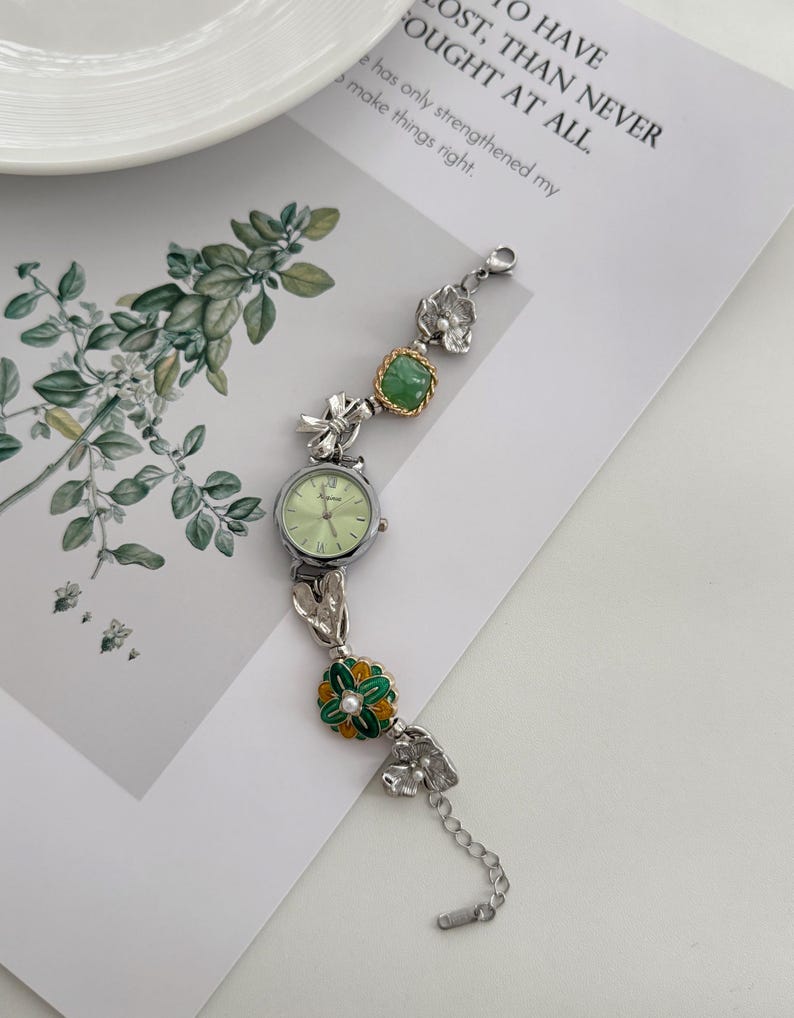 May include: A silver-toned wristwatch with a light green face and a decorative band. The band features floral and leaf accents, including a green and gold flower with a pearl center. The watch is on a white surface with text in the background.