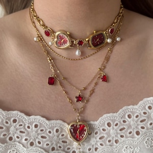 Pode incluir: Colar dourado em camadas com mostradores de relógio vermelhos, pingentes em forma de coração e detalhes de pedras vermelhas. O colar inclui pérolas e um berloque em forma de estrela. Os mostradores mostram as horas.