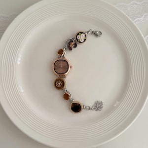 K&ouml;nnte beinhalten: Eine ros&eacute;goldfarbene Uhr mit quadratischem Zifferblatt und dekorativem Armband. Das Armband hat florale und edelsteinartige Akzente in Braun-, Gold- und Silbert&ouml;nen. Die Uhr wird auf einem wei&szlig;en Teller pr&auml;sentiert.