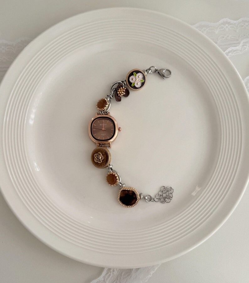 K&ouml;nnte beinhalten: Eine ros&eacute;goldfarbene Uhr mit quadratischem Zifferblatt und dekorativem Armband. Das Armband hat florale und edelsteinartige Akzente in Braun-, Gold- und Silbert&ouml;nen. Die Uhr wird auf einem wei&szlig;en Teller pr&auml;sentiert.