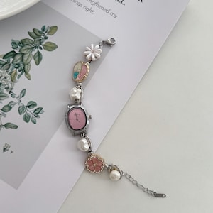 May include: A silver-toned bracelet watch with a pink face and decorative floral and pearl accents. The watch features a small, oval-shaped face and is adorned with white and pink flowers and faux pearls. The text on the paper reads: "THAN NEVER OUGHT AT ALL."