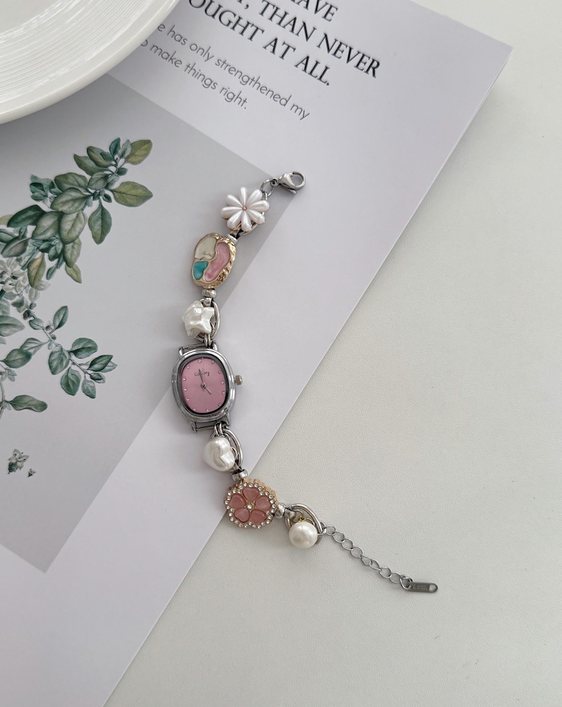 May include: A silver-toned bracelet watch with a pink face and decorative floral and pearl accents. The watch features a small, oval-shaped face and is adorned with white and pink flowers and faux pearls. The text on the paper reads: "THAN NEVER OUGHT AT ALL."