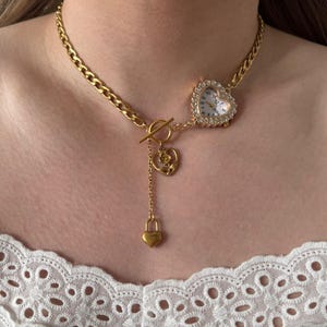 May include: Gold-toned necklace featuring a heart-shaped watch with a crystal-studded bezel. The necklace has a chain with a toggle clasp and a dangling heart pendant with a lock charm. The watch face displays the time.