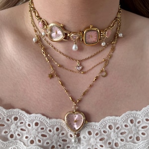 Pode incluir: Um colar dourado de várias camadas com mostradores de relógio e berloques. A camada superior é um gargantilha com dois mostradores de relógio, um em forma de coração e outro quadrado, com mostradores rosa. Camadas adicionais incluem berloques de coração, lua e estrela, e pérolas falsas.