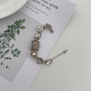May include: A silver-toned watch with a rectangular face and a decorative bracelet. The bracelet features various embellishments, including clear crystals, pearls, and floral designs. The watch is on a white surface with text that reads "TO HAVE LOST, THAN NEVER FOUGHT AT ALL."