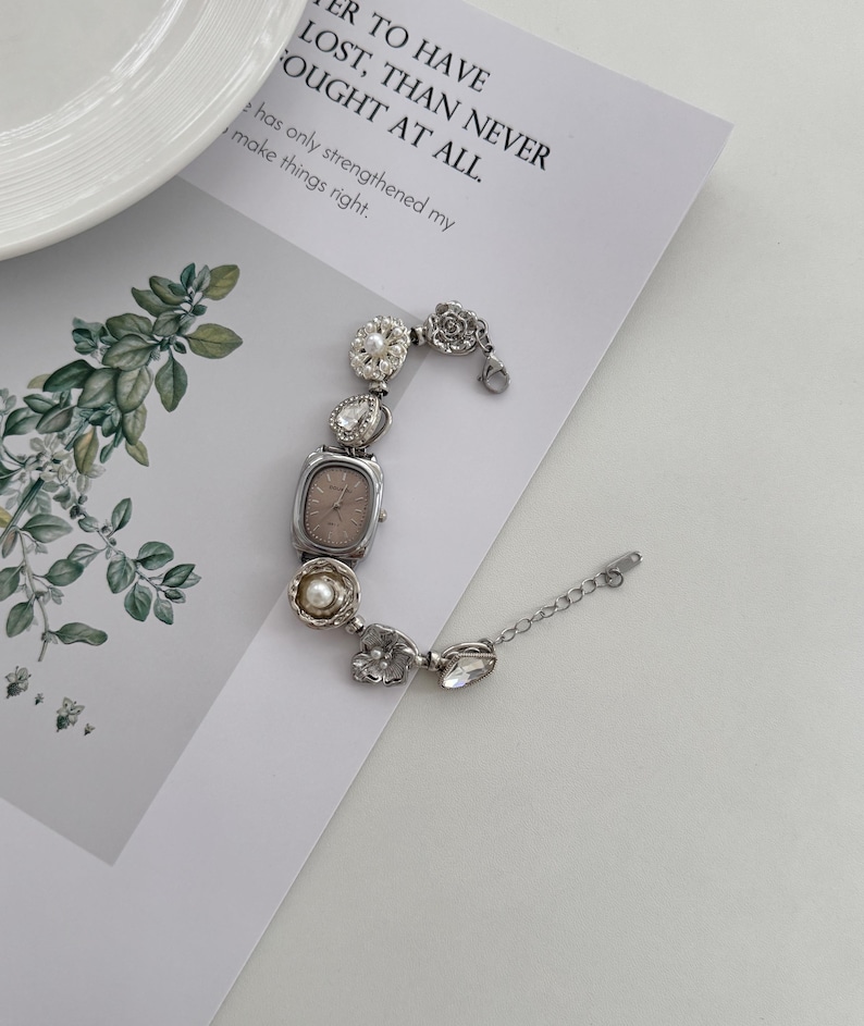 May include: A silver-toned watch with a rectangular face and a decorative bracelet. The bracelet features various embellishments, including clear crystals, pearls, and floral designs. The watch is on a white surface with text that reads "TO HAVE LOST, THAN NEVER FOUGHT AT ALL."