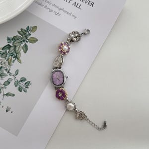 May include: A silver-toned bracelet watch with a rectangular lavender face and decorative floral and pearl accents. The watch is adorned with purple and white flowers and a delicate chain. The text on the paper reads: "R TO HAVE LOST, THAN NEVER FOUGHT AT ALL."