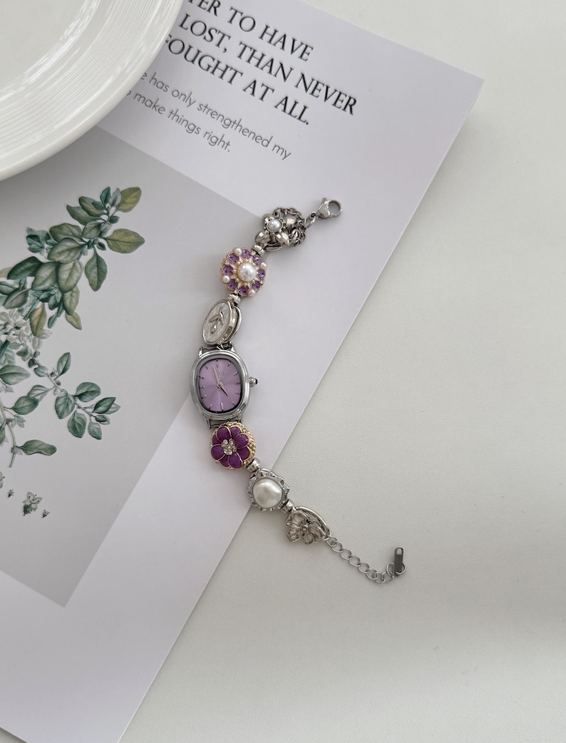 May include: A silver-toned bracelet watch with a rectangular lavender face and decorative floral and pearl accents. The watch is adorned with purple and white flowers and a delicate chain. The text on the paper reads: "R TO HAVE LOST, THAN NEVER FOUGHT AT ALL."