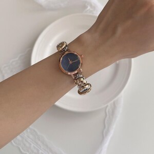 K&ouml;nnte beinhalten: Eine ros&eacute;goldene Armbanduhr mit dunkelblauem Zifferblatt und dekorativem Armband. Das Armband hat goldene und silberne Akzente. Die Uhr wird an einem Handgelenk vor wei&szlig;em Hintergrund gezeigt.