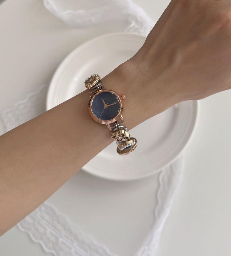 K&ouml;nnte beinhalten: Eine ros&eacute;goldene Armbanduhr mit dunkelblauem Zifferblatt und dekorativem Armband. Das Armband hat goldene und silberne Akzente. Die Uhr wird an einem Handgelenk vor wei&szlig;em Hintergrund gezeigt.