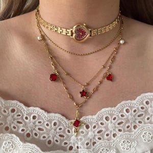 Può includere: Set di collane dorate a strati con un orologio a forma di cuore, dettagli di perle e vari ciondoli. I ciondoli includono cuori, stelle e quadrati rossi, e un ciondolo a croce con una gemma rossa.
