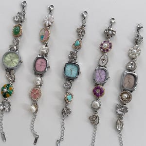 May include: Five silver-toned wristwatches with decorative embellishments. Each watch features a different colored face, including green, pink, and blue. The bands are adorned with pearls, flowers, and other decorative elements.