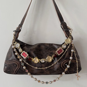 May include: A dark brown faux leather shoulder bag adorned with a decorative chain assembly featuring multiple gold-tone wristwatches, red rectangular watch faces, white faux pearls, and colorful crystal accents, including a dangling cross charm.