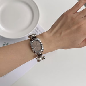 May include: Silver-toned rectangular watch with a light-colored face. The watch band features floral and pearl accents. The watch is worn on a wrist, showcasing its design and style. A classic accessory.