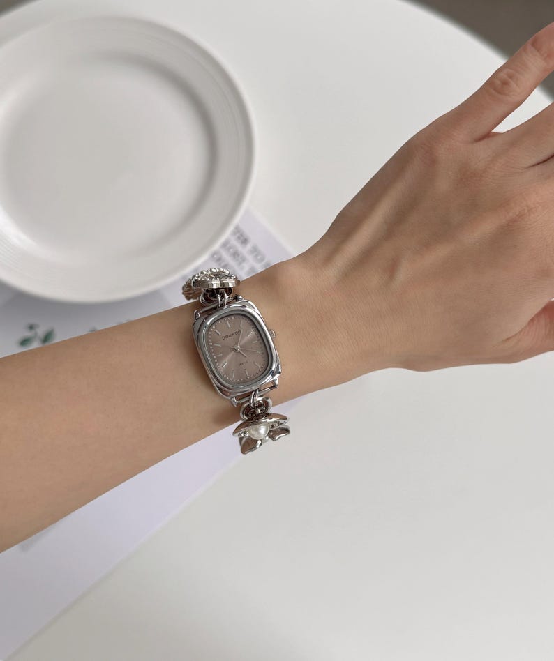 May include: Silver-toned rectangular watch with a light-colored face. The watch band features floral and pearl accents. The watch is worn on a wrist, showcasing its design and style. A classic accessory.