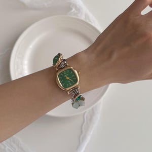 K&ouml;nnte beinhalten: Eine goldfarbene Armbanduhr mit einem quadratischen gr&uuml;nen Zifferblatt und goldenen Zeigern. Das Uhrenarmband ist eine Mischung aus goldenen und silbernen Gliedern, verziert mit gr&uuml;nen Steinakzenten und einem kleinen Blumenanh&auml;nger. Die Uhr befindet sich an einem Handgelenk.