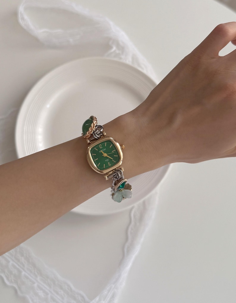 K&ouml;nnte beinhalten: Eine goldfarbene Armbanduhr mit einem quadratischen gr&uuml;nen Zifferblatt und goldenen Zeigern. Das Uhrenarmband ist eine Mischung aus goldenen und silbernen Gliedern, verziert mit gr&uuml;nen Steinakzenten und einem kleinen Blumenanh&auml;nger. Die Uhr befindet sich an einem Handgelenk.