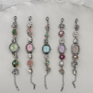 Puede incluir: Cinco relojes plateados con adornos decorativos. Cada reloj tiene una esfera de diferente color: verde, rosa, azul, morado y beige. Las correas de los relojes presentan varios abalorios, incluyendo flores, perlas y piedras preciosas. Cada reloj tiene una cadena al final.