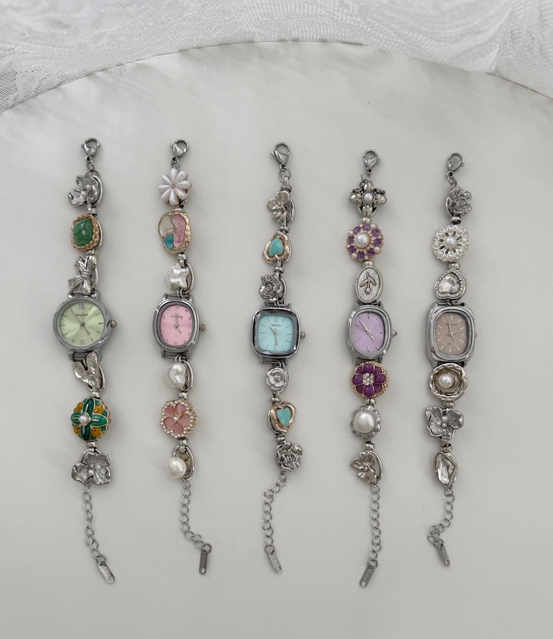 May include: Five silver-toned watches with decorative embellishments. Each watch has a different colored face: green, pink, blue, purple, and tan. The watch bands feature various charms, including flowers, pearls, and gemstones. Each watch has a chain at the end.