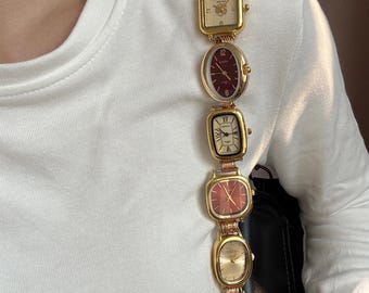 May include: A gold-toned watch with multiple watch faces of varying shapes, including oval, square, and rectangular. The watch faces have burgundy and cream-colored dials. The watch is connected by a gold chain.