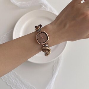 K&ouml;nnte beinhalten: Ros&eacute;goldene Armbanduhr mit quadratischem Zifferblatt und braunem Zifferblatt. Das Uhrenarmband hat dekorative Charms und wird am Handgelenk getragen. Die Uhr wird auf einer wei&szlig;en Oberfl&auml;che mit einem wei&szlig;en Teller und Spitze pr&auml;sentiert.