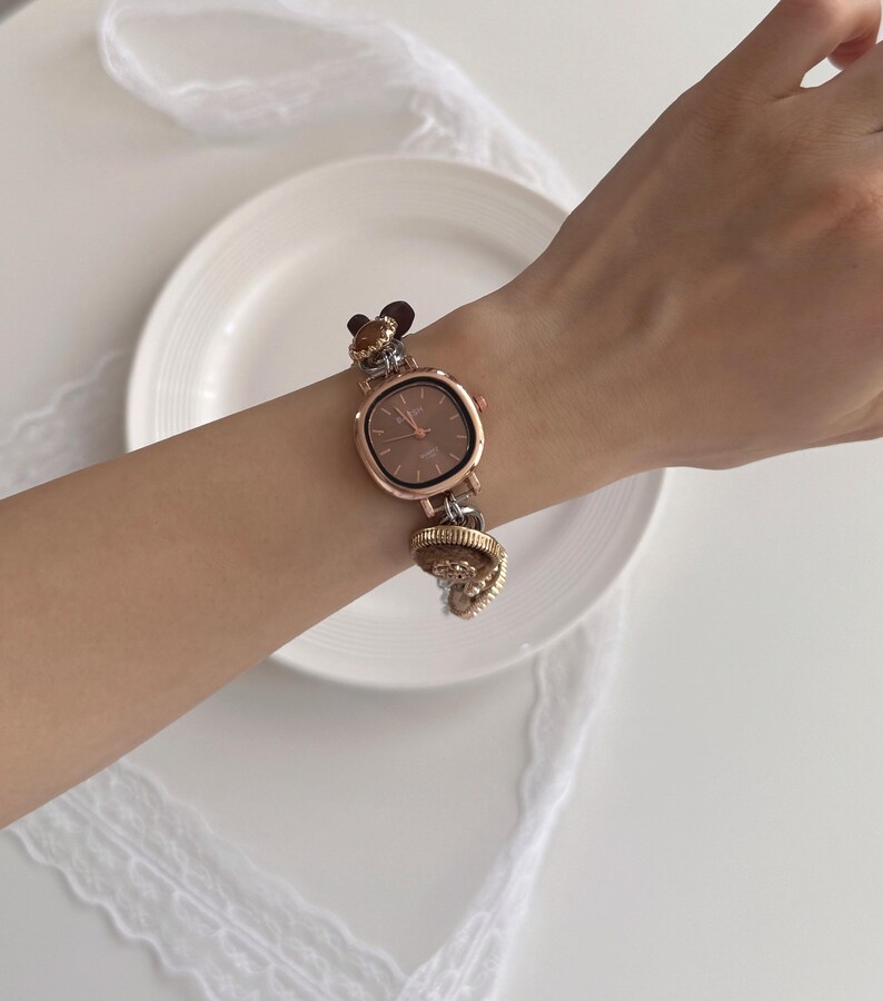 K&ouml;nnte beinhalten: Ros&eacute;goldene Armbanduhr mit quadratischem Zifferblatt und braunem Zifferblatt. Das Uhrenarmband hat dekorative Charms und wird am Handgelenk getragen. Die Uhr wird auf einer wei&szlig;en Oberfl&auml;che mit einem wei&szlig;en Teller und Spitze pr&auml;sentiert.