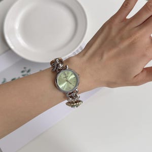 May include: Silver-toned wristwatch with a light green face and Roman numeral hour markers. The watch has a decorative band with ornate details. The watch is displayed on a wrist.