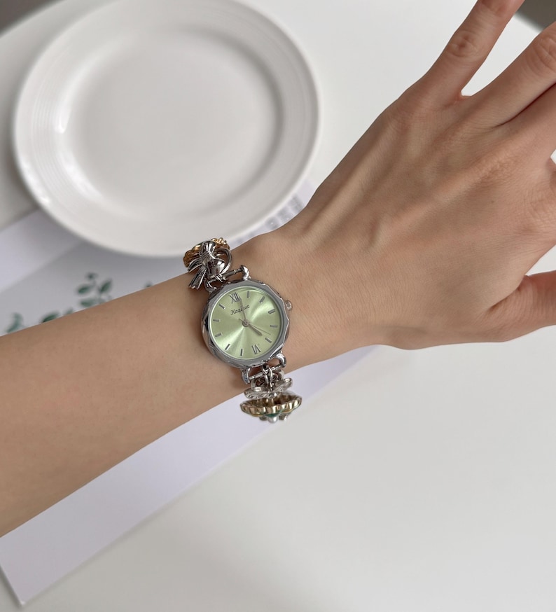 May include: Silver-toned wristwatch with a light green face and Roman numeral hour markers. The watch has a decorative band with ornate details. The watch is displayed on a wrist.