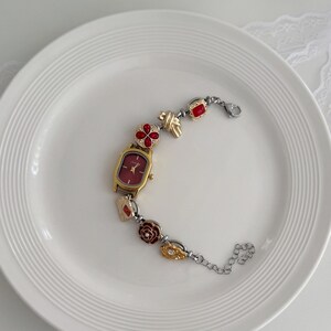 K&ouml;nnte beinhalten: Eine goldfarbene Armbanduhr mit rechteckigem Zifferblatt und burgunderfarbenem Zifferblatt. Das Armband hat dekorative Elemente, darunter rote bl&uuml;tenf&ouml;rmige Akzente, goldene Knoten und rote quadratische Edelsteine. Die Uhr liegt auf einem wei&szlig;en Teller.