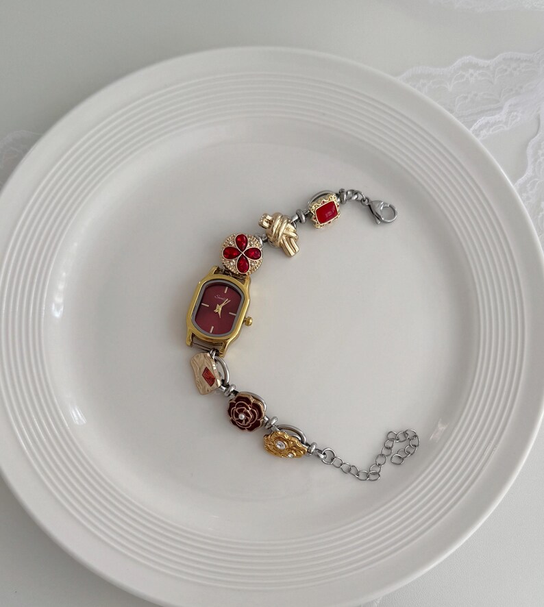 K&ouml;nnte beinhalten: Eine goldfarbene Armbanduhr mit rechteckigem Zifferblatt und burgunderfarbenem Zifferblatt. Das Armband hat dekorative Elemente, darunter rote bl&uuml;tenf&ouml;rmige Akzente, goldene Knoten und rote quadratische Edelsteine. Die Uhr liegt auf einem wei&szlig;en Teller.