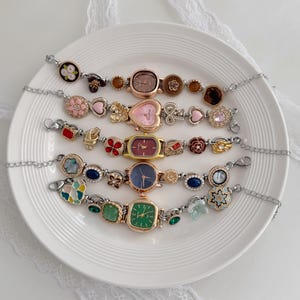 K&ouml;nnte beinhalten: Eine Sammlung dekorativer Armbanduhren mit verschiedenen Zifferblattformen und Verzierungen. Die Uhren haben verschiedenfarbige Zifferbl&auml;tter, darunter rosa, gr&uuml;n und blau, und sind mit floralen und edelsteinartigen Akzenten verziert. Die Armb&auml;nder sind silberfarben.