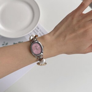 May include: Silver-toned wristwatch with a pink oval face and silver hands. The watch has a silver band with decorative pearl-like accents. The watch is on a wrist.