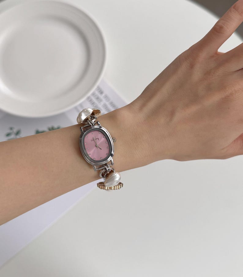 May include: Silver-toned wristwatch with a pink oval face and silver hands. The watch has a silver band with decorative pearl-like accents. The watch is on a wrist.