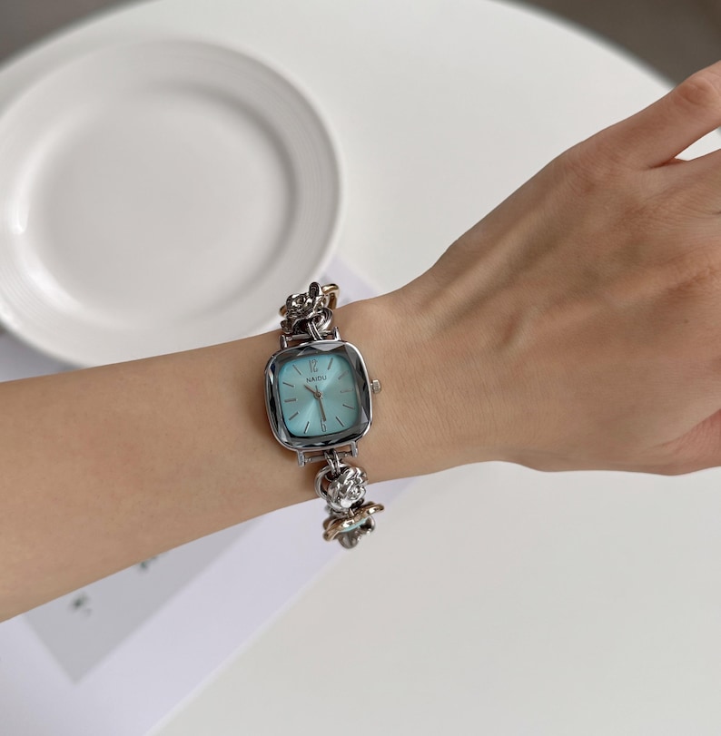 May include: A silver-toned wristwatch with a square face and a light blue dial. The watch has a decorative chain-link band with floral accents. The watch is on a wrist, and the background includes a white plate and a white surface.