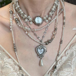 Puede incluir: Un collar de varias capas con perlas blancas, cuentas de vidrio transparente y cuentas metálicas plateadas. La pieza incluye una esfera de reloj redonda en la clavícula y un colgante de reloj en forma de corazón con una perla en forma de lágrima.
