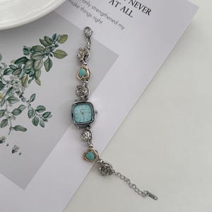 May include: A silver-toned bracelet watch with a square, light blue face. The watch band features decorative silver and gold-toned floral and heart-shaped accents with turquoise stones. The text on the paper reads: "R TO HAVE LOST, THAN NEVER OUGHT AT ALL."