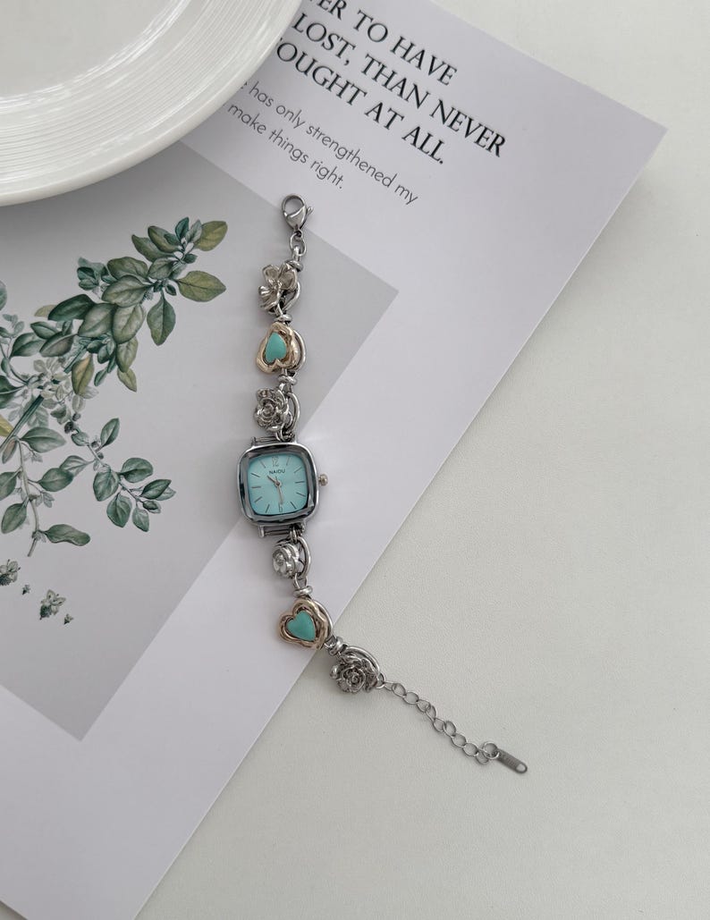 May include: A silver-toned bracelet watch with a square, light blue face. The watch band features decorative silver and gold-toned floral and heart-shaped accents with turquoise stones. The text on the paper reads: "R TO HAVE LOST, THAN NEVER OUGHT AT ALL."