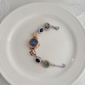 K&ouml;nnte beinhalten: Ein ros&eacute;goldenes Armband mit einem marineblauen Zifferblatt und dekorativen blauen und goldenen Akzenten. Das Armband verf&uuml;gt &uuml;ber eine Reihe von runden und ovalen Verzierungen mit verschiedenen Farben und Texturen, verbunden durch eine silberne Kette.