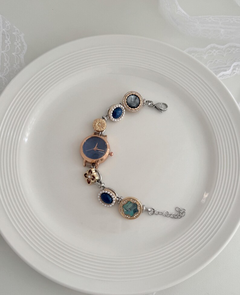 K&ouml;nnte beinhalten: Ein ros&eacute;goldenes Armband mit einem marineblauen Zifferblatt und dekorativen blauen und goldenen Akzenten. Das Armband verf&uuml;gt &uuml;ber eine Reihe von runden und ovalen Verzierungen mit verschiedenen Farben und Texturen, verbunden durch eine silberne Kette.