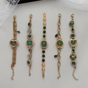 May include: Five gold-coloured wristwatches with green watch faces and decorative green gemstones. The watch faces have different shapes, including a heart and oval. The watch bands are also adorned with green gems.