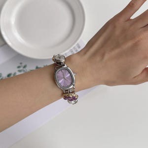 May include: A silver-toned wristwatch with a light purple face and a decorative band featuring flower-shaped charms. The watch has an oval face and is worn on a wrist. A white plate is in the background.