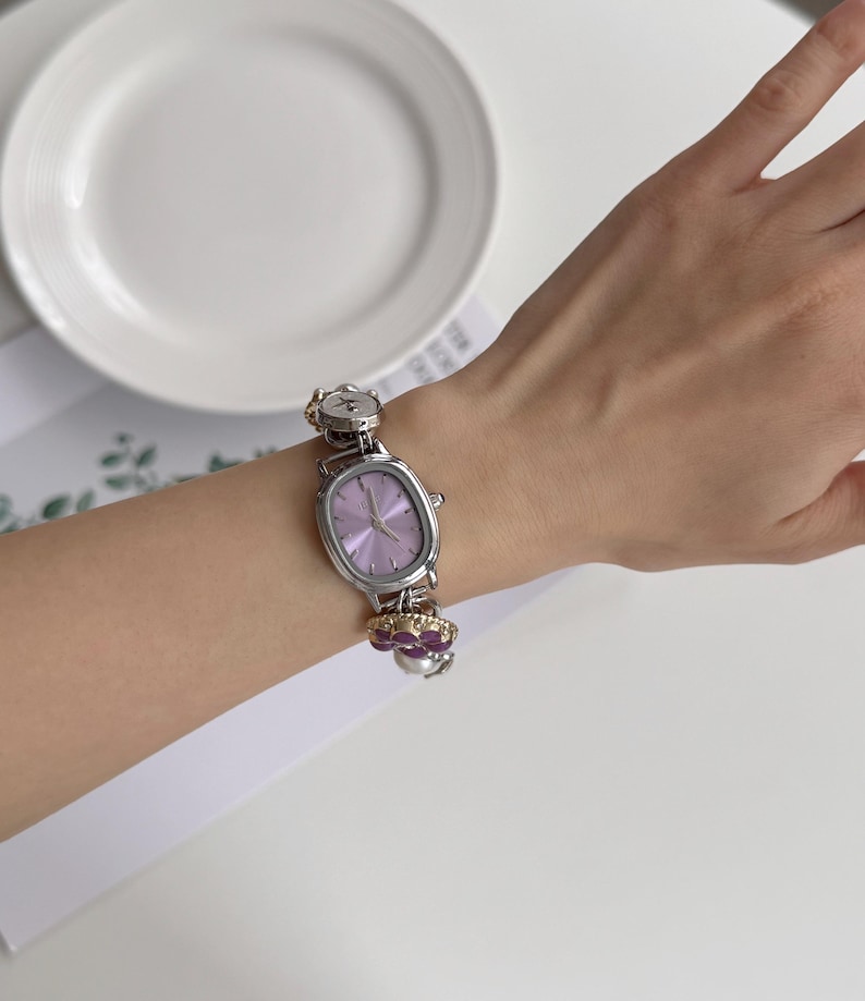 May include: A silver-toned wristwatch with a light purple face and a decorative band featuring flower-shaped charms. The watch has an oval face and is worn on a wrist. A white plate is in the background.