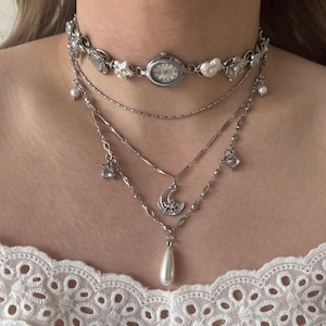 Può includere: Set di collane d'argento multistrato. Lo strato superiore è un girocollo con quadrante e dettagli floreali. Sotto ci sono delicate catene con pendenti di perle e ciondoli, tra cui un design a luna e stella e una perla a goccia.