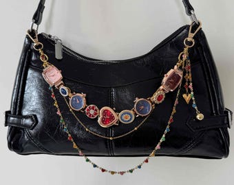 May include: Black shoulder bag with a decorative chain of colorful watches, hearts, and beads. The bag has a shiny finish and a silver zipper. The chain is attached to the bag with gold-colored clasps. The bag's strap is black.