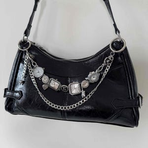 Könnte beinhalten: Schwarze Schultertasche mit silberfarbener Hardware und einer dekorativen Kette aus Uhren im Vintage-Stil. Die Tasche hat eine glänzende Oberfläche und einen Reißverschluss. Der Riemen ist verstellbar.