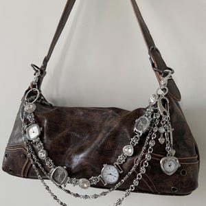 Könnte beinhalten: Eine dunkelbraune Umhängetasche aus Leder in Used-Optik mit einer dekorativen silberfarbenen Kette, die über die Vorderseite drapiert ist. Die Kette ist mit mehreren kleinen Armbanduhren, herzförmigen Anhängern und klaren Kristallen verziert.