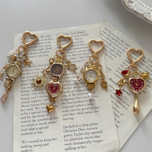 May include: Four gold-toned watch charms with heart-shaped clasps and decorative dangles. Each charm features a small watch face, some with heart-shaped dials, and various embellishments like pearls and flowers. The charms rest on a vintage paper background.