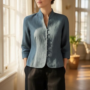May include: A blue linen jacket with a mandarin collar and three-quarter sleeves. The jacket has a button-down front and is paired with black pants. The outfit is suitable for a professional or casual setting.