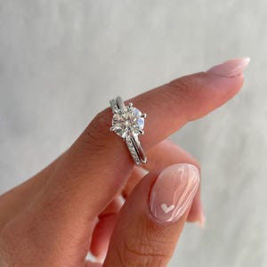 Könnte beinhalten: Ein silberner Verlobungsring mit einem großen, runden, klaren Edelstein. Der Ring wird von einem Finger mit hellrosa Nagellack und einem kleinen weißen Herzdesign gehalten.